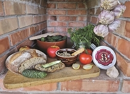 Drewniana deska, a na niej ułożony chleb, pojemnik ze smalcem, ogórki, pęczek kopru, pomidor, jabłko i przekorjona na pół cebula