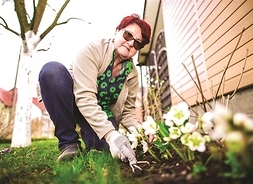 kobieta pracuje w ogródku, pochyla się nad kwiatami i pozuje do zdjęcia, w tle drzewo, obok ściana budynku