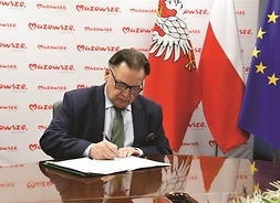 Marszałek Adam Struzik podczas podpisywania dokumentu. Zdjęcie na tle flag oraz banneru Mazowsze. serce Polski.