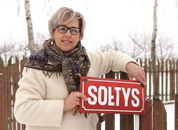 Kobieta w zimowej kurtce z chustą na plecach stoi przy plocie, trzymając metalową tabliczkę z napisem Sołts.