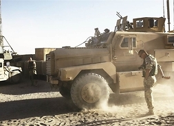 Żołnierz biorący udział w misji w Afganistanie stoi przy pojeździe wojskowym. W tle koleny pojazd i żołnierz stojący przy metalowym maszcie..