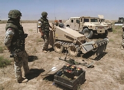 Dwaj żołnierze w pełnym umundurowaniu i ochronnych hełmach stoją przy robotach oczyszczających pola z min.