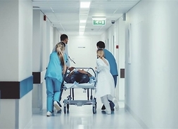 Personel medyczny transortuje po szpitalnym korytarzu chorego leżącego na łóżku medycznym.