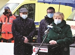 przed mikrofonem stoją kobieta i dwóch mężczyzn, w tle ratownik medyczny i karetka