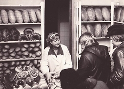 Zdjęcie archiwalne. Wnętrze piekarni z regałami z pieczywem. Kobieta stojąca za ladą, przed którą ustawiła się kolejka kupujących.