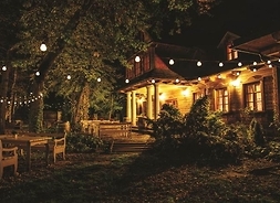 Widok z boku na front drewnianego dworku, który jest rożświetlony wiszącymi lampkami. Zdjęcie robione nocą