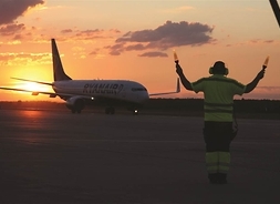 Pracownik lotniska za pomocą dwóch flar trzymanych w uniesionych nad głową dłoniach daje sygnały pilotowi samolotu pasażerskiego, który porusza sę po płycie lotniska.