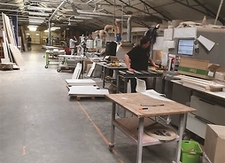 Hala produkcyjna zakładu meblarskiego, na zdjęciu widoczne są płyty przygotowane do produkcji mebli, urządzenia oraz pracownicy, podczas wykonywania swoich zajęć.