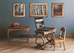 Drewniany stolik, na który stoi otwarty laptop. Przy stoliku stoi drewniane krzesło, za stolikiem drewniana konsola. Na ścianie wiszą trzy obrazy w drewnanych ramach, przedstawiające naturę.