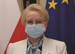 na tle flag Polski i Unii Europejskiej stoi elegancka kobieta w maseczce, trzyma teczkę z wyróżnieniem
