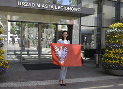 kobieta w jasnej marynarce i spodniach stoli przed wejściem do leionowskiego ratusza, trzyma przed sobą rozłożoną flagę Mazowsza