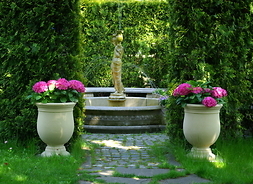 kamienna fotalna  stoi na środku, obok dwie donice z różowymi kwiatami, w tle zielony ogród