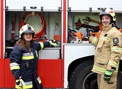 Strażak i strażaczka na tle otwartego wozu strażackiego.