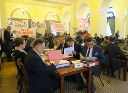 Widok na salę obrad. Wokół radnych, siedzących przy stołach stoją protestujący mieszkańcy Mazowsza