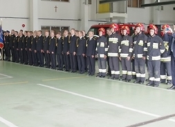 Stojący w szeregu strażacy, ubrani w stroje służbowe albo jak na akcję