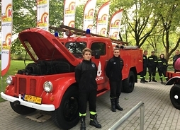 samochód strażacki ma podniesioną maskę obok stoją dwaj młodzi strażacy