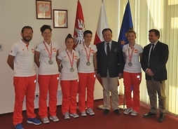 Sportowcy w dresach biało-czerwonych z samorządowcami na tle flag Polski, UE i Mazowsza