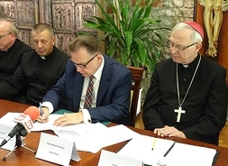 marszałek w trakcie podpisywania po jego lewej stronie siedzi biskup płocki