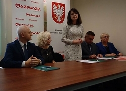 Przy stole siedzą beneficjenci z powiatu płońskiego, przemawia Janina Ewa Orzełowska