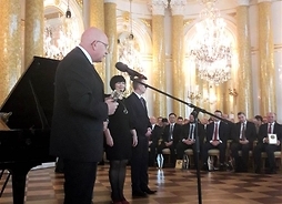 Samorządowiec przemawia przed mikrofonem w sali Zamku Królewskiego, w tle siedzą uczestnicy uroczystości i stoi fortepian
