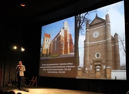 Historyk stoi przed ekranem kinowym na którym wyświetlane jest zdjęcie katedry w Łomży