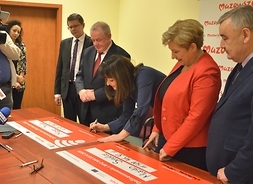 Wicemarszałek Janina Ewa Orzełowska składa podpis na pamiątkowym czeku. Obok stoją członek zarządu Elżbieta Lanc, beneficjent oraz dziennikarze