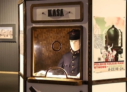 Manekin w stroju kolejarskim siedzący w okienku kasowym, obok plakat z rysunkiem pociągu i podróżnych