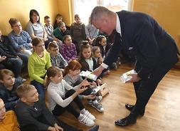 grupa najmłodszych uczniów siedzi na sali gimnastycznej; zestawy odblakowe ręcza im członek zarządu Rafał Rajkowski