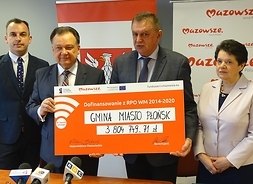 cztery osoby stoją, 2 z nich trzymają w ręku symboliczny duży czek z wpisaną kwotą dofinansowania dla Płońska