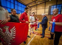 Dwóch wręczających cztery osoby oczekują na odbiór medali i trzymają w rękach flagę Mazowsza