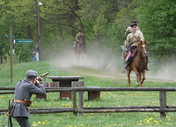 Na pierwszym planie żołnierz celuje ze strzelby w nadjeżdżającego na koniu jeźdźca