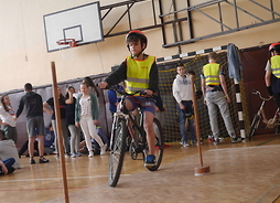 Na pierwszym planie znajduje się uczeń na rowerze pomiędzy pachołkami. W tle widać uczniów zgromadzonych na sali gimnastycznej