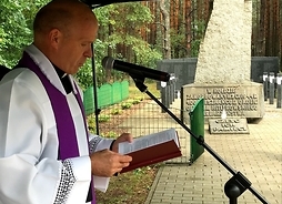 Ksiądz czyta, w tle pomnik  hołdzie zamordowanym ofiarom hitlerowskiego barbarzyństwa