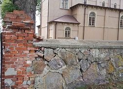 Zniszczony kamienny parkan, w tle drewniany kościół