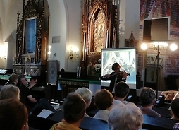 Na zdjęciu widać artystę grającego na skrzypcach i zasłuchaną publiczność