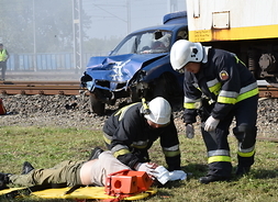 Dwóch strażaków stoi pochylonych nad poszkodowanym kierowcą pojazdu, który zderzył się z pociągiem