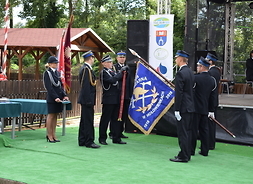 Trzech strażaków stoi na zielonym dywanie przed sceną prezentując sztandar. Przed nimi stoi komendant i nakłada na sztandar czerwoną wstęgę