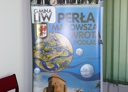 Na zdjęciu baner promujący gminę Liw. Na banerze jest Orkiestra Dęta, Zamek w Liwie i meandrujący Liwiec.