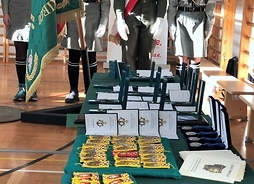Na długim stole leżą odznaki i dyplomy dla wyróżnionych. Obok stołu widać harcerzy i chorągiew