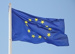 niebieska flaga Unii Europejskiej powiewająca na maszcie. W tle błękitne niebo