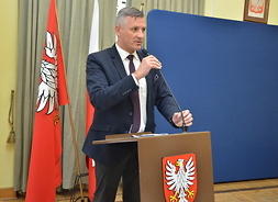 Przy mównicy stoi wicemarszałek Rafał Rajkowski. Jedną ręką podtrzymuje mikrofon, w tle flagi UE, Polski i Mazowsza