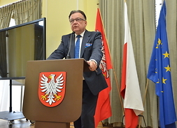 Marszałek Struzik przemawia przy mównicy. Widać mównicę z godłem województwa, w tle flagi UE, Polski i Mazowsza
