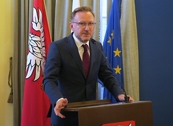 Przy mównicy stoi mężczyna w garniturze i okularach. W tle flagi UE, Polski i Mazowsza