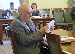 Na krześle, ustawiony bokiem, siedzi starszy mężczyna w okularach trzyma w uniesionych rękach dokument, który czyta. W tle widać krzesła i dwóch radnych sejmiku
