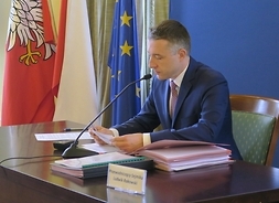 Przy stole, na którym leżą dokumenty, siedzi mężczyzna w garniturze i mówi do mikrofonu. W tle flagi UE, Polski i Mazowsza