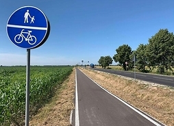 Widok na asfaltową ścieżkę pieszo-rowerową, która jest przy drodze. na pierwszym planie znak drogowy z oznaczeniem trasy pieszo-rowerowej