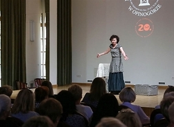 Zdjęcie przedstawia aktorkę Joannę Szczepkowską podczas występu na scenie oraz siedzącą publiczność