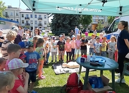 Duża grupa dzieci w słoneczny dzień stoi wokół pani z mikrofonem. Obok stoi stolik z przedmiotami pierwszej pomocy