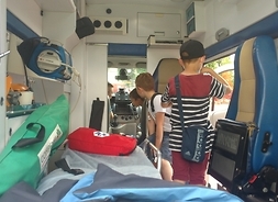 Wnętrze ambulansu ratowniczego. W środku jest trzech chłopców. Jeden stoi tyłem przy łóżku i patrzy na dwóch, którzy siedzą za kierownicą.