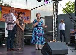 Członek zarządu Elżbieta Lanc stoi na scenie. W prawym ręku trzyma mikrofon. Obok niej po lewej stronie widać mężczyznę. po prawej - kobietę i mężczyznę stojących blisko siebie